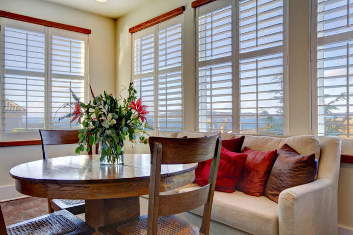 Salón con cortinas venecianas de madera blancas mesa y sillas de madera marrón sofá claro con cojines rojos